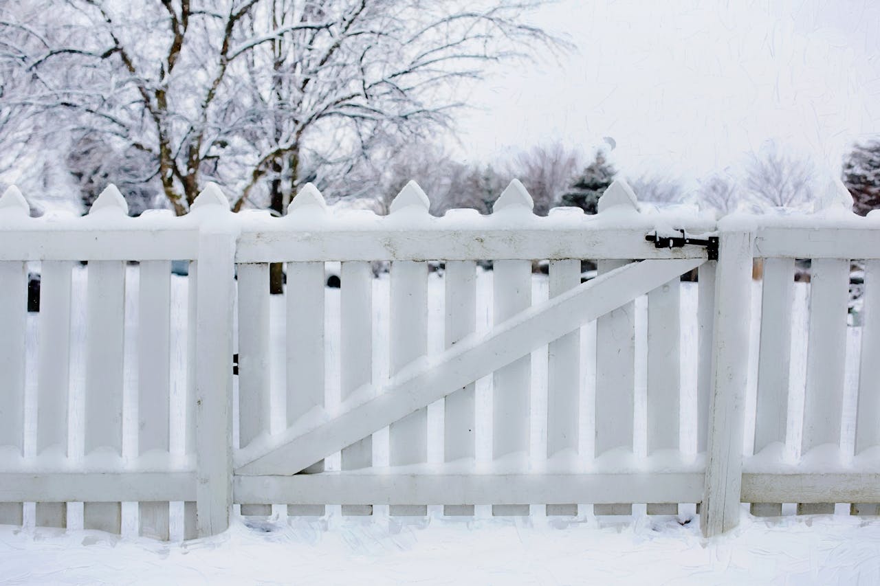 A snow-covered white picket fence in a serene winter landscape, surrounded by frosted trees.
